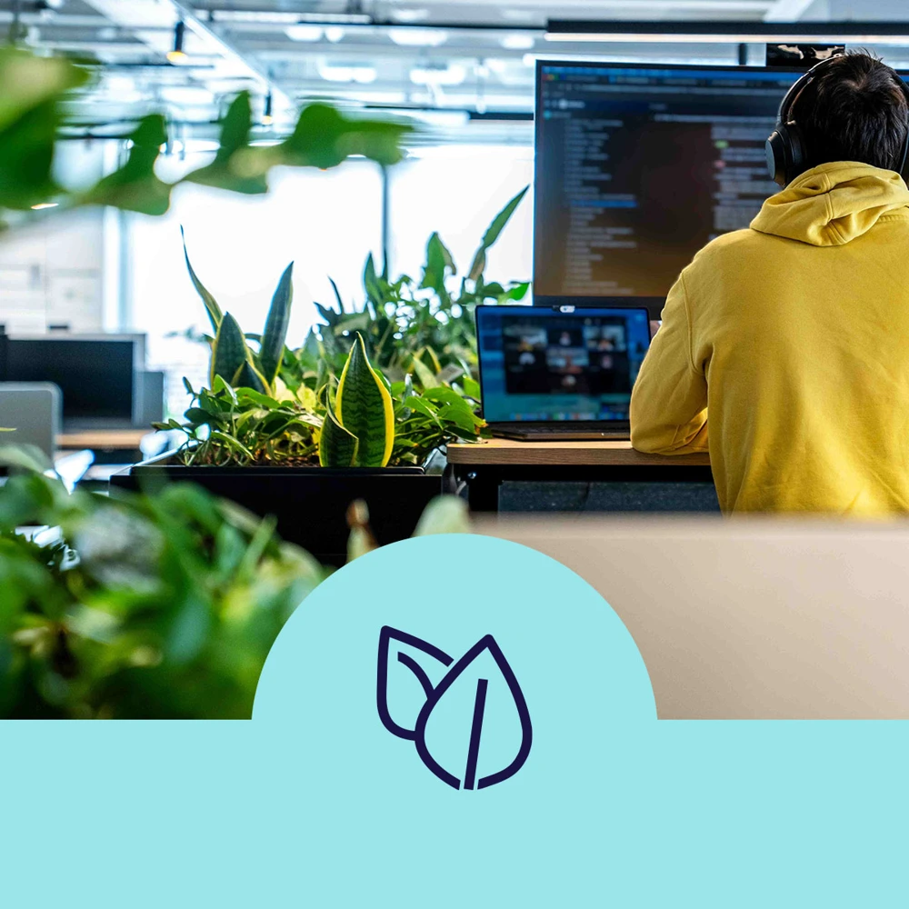 A person in a yellow hoodie is seated at a workstation with multiple screens, surrounded by green plants. The environment appears to be an open office space. A blue graphic with leaf icons is overlaid at the bottom of the image.
