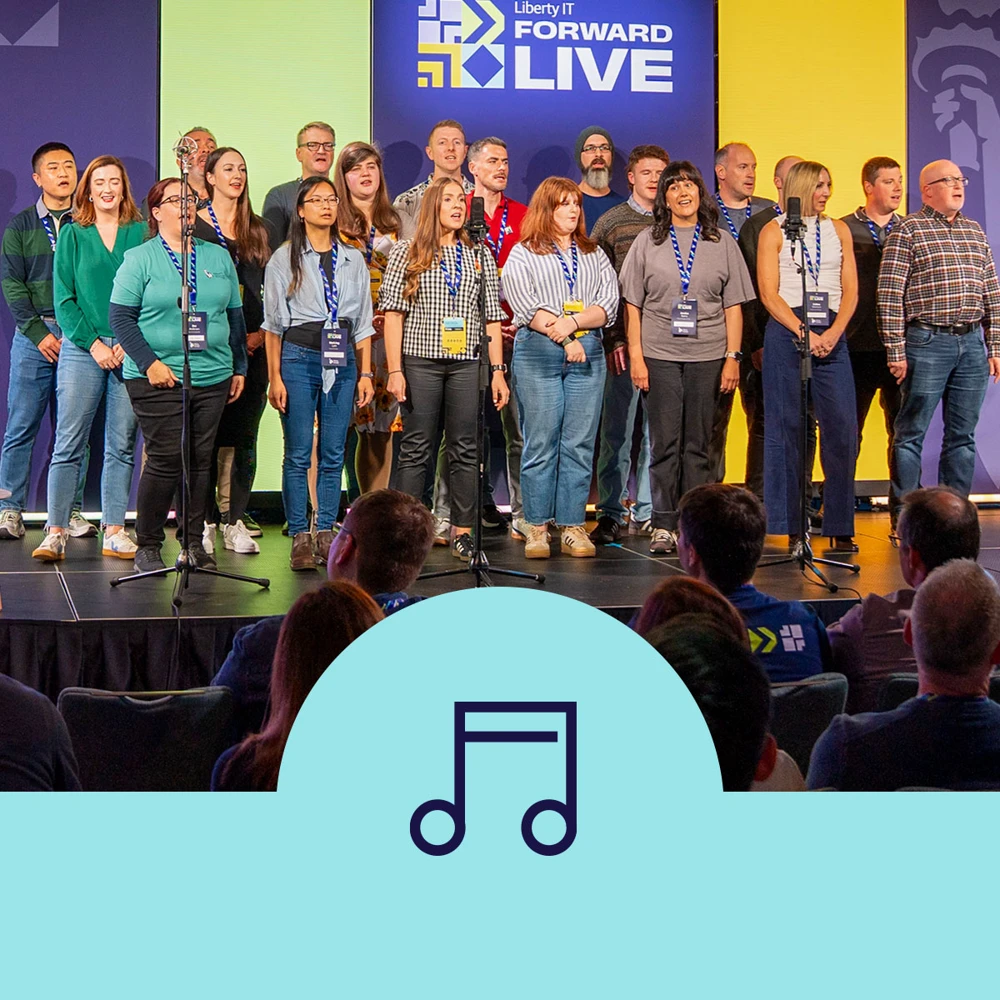 A group of people stands on a stage wearing conference badges, with a "Liberty IT Forward Live" banner behind them. A seated audience is in the foreground.