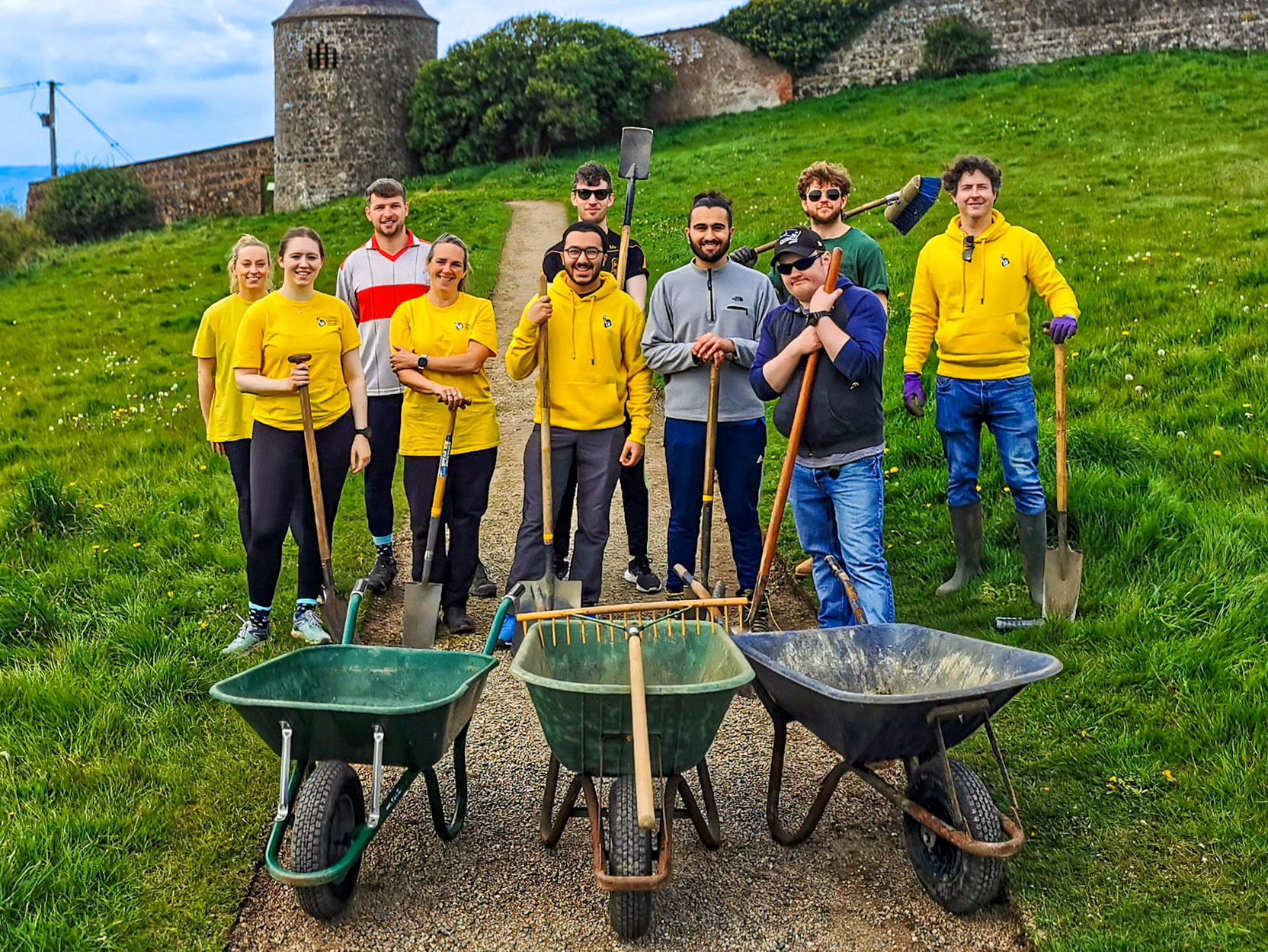 A group of people pose outdoors with three wheelbarrows, rakes, and shovels on a gravel path. They are in front of a green field and an old stone tower.