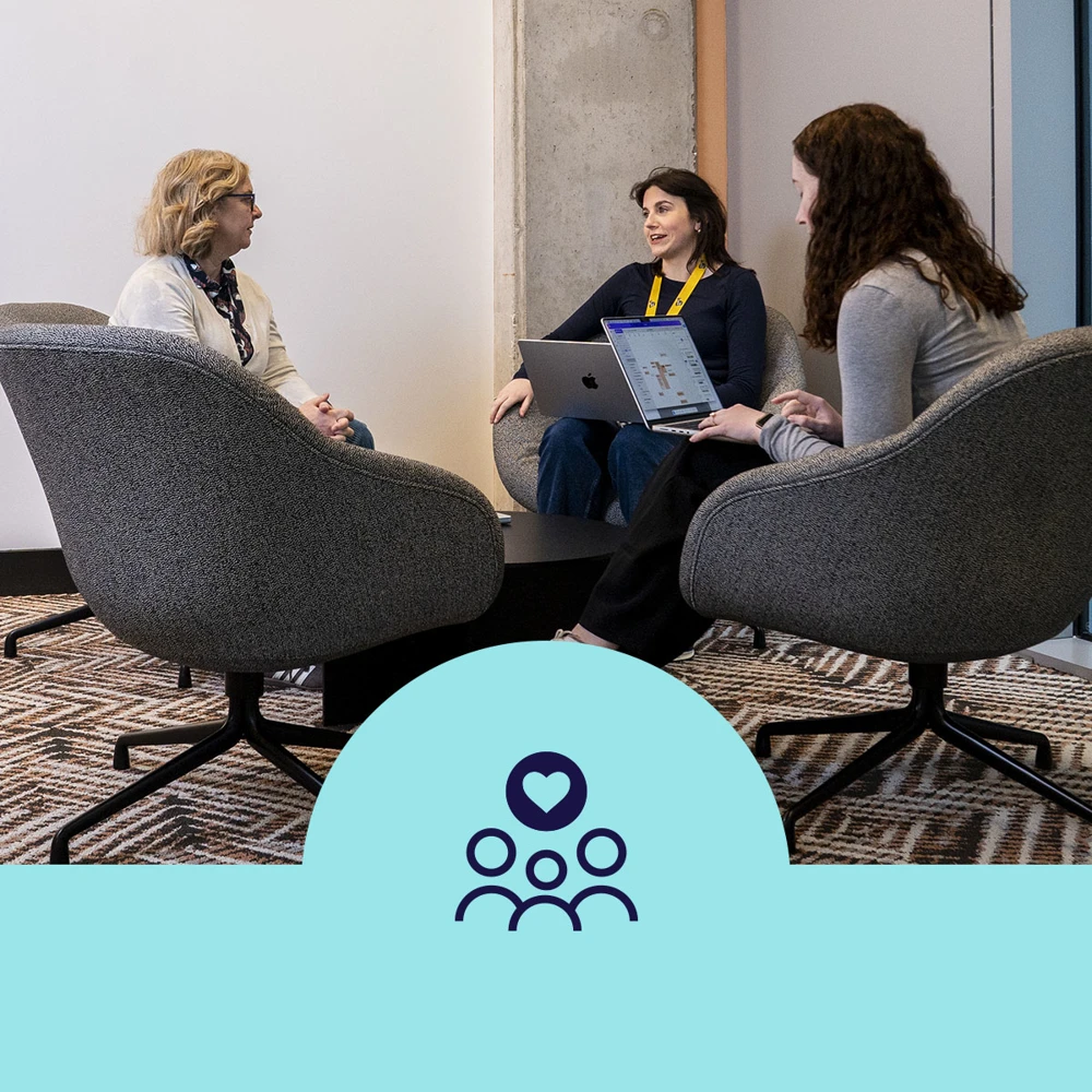 A group of three people seated in gray chairs having a discussion. Two individuals are holding open laptops. The background has a white wall and a patterned carpet. A light blue banner with an icon of three people and a heart is at the bottom of the image.
