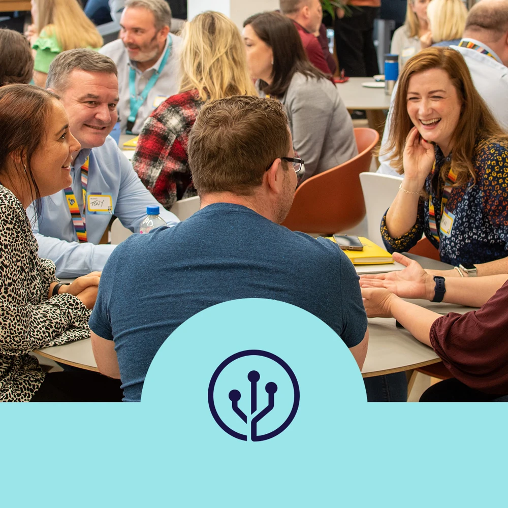 A group of people are sitting at tables, engaged in conversation and smiling, inside a busy room. Various individuals are wearing name tags, and several have open notebooks and laptops on the table. The foreground features a blue semicircle with a technology-inspired logo.