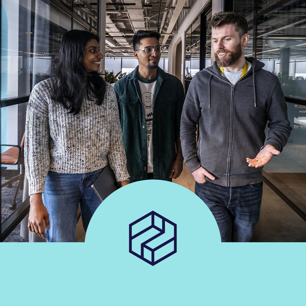 Three people walking down an office hallway, engaged in conversation. They wear casual attire. A logo is displayed below, resembling an intertwined geometric shape.